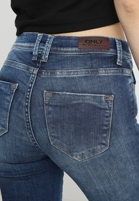 Wąskie niebieskie jeansy z wysokim stanem, z brązową skórzaną łatą z tyłu i dwoma tylniami kieszeniami. Pranie wzdłuż ud.