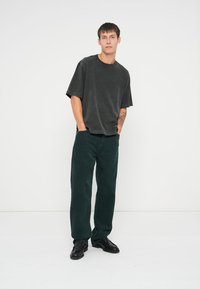 Mörkgrå oversized t-shirt med korta ärmar, i kombination med enfärgade gröna kordbyxor. Svarta läderskor, stående mot en enkel bakgrund.