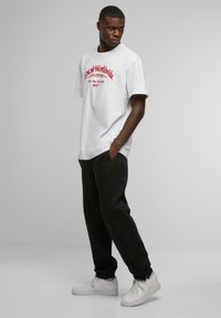 White cotton t-shirt with red graphic text, paired with black sweatpants and white sneakers. Model standing with hands in pockets.