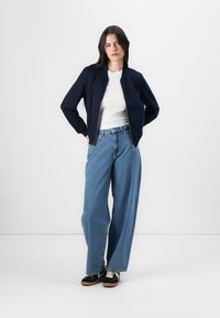 Navy bomber jacket with a zipper, white crew neck shirt, and wide-leg blue denim jeans. Black and white sneakers complement the outfit.