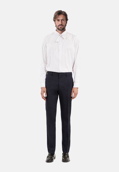 Wit gestreepte shirt met een strikje, gecombineerd met donkerblauwe op maat gemaakte broeken. Model draagt zwarte schoenen en staat tegen een neutrale achtergrond.