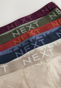Pila de ropa interior doblada con cinturillas elásticas en burdeos, verde oliva, óxido, azul, azul marino y beige, cada una con la palabra "NEXT" en relieve.
