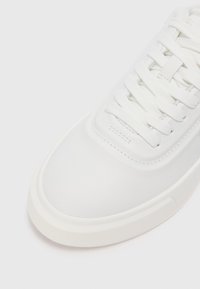 Adidași albi cu un superior sintetic neted, vârf rotund și un design cu șireturi plate. Talpa este ușor ridicată, fără modele vizibile.