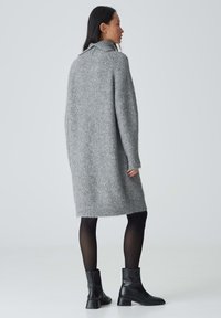 Grau gestricktes Sweatkleid mit hohem Kragen, langen Ärmeln und geradem Schnitt. Kombiniert mit schwarzen Stiefeletten und durchsichtigen schwarzen Leggings.