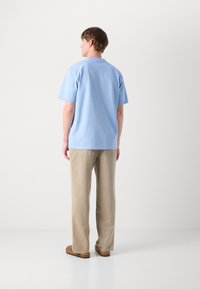 Ανοιχτό μπλε μπλουζάκι κοντού μανικιού από βαμβάκι, φορεμένο με μπεζ παντελόνι χαλαρής γραμμής και καφέ μοκέτες, δείχνοντας έναν απλό, casual σχεδιασμό.