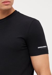 Camiseta negra de manga corta hecha de una tela suave. Cuenta con un cuello redondo y un detalle de logo blanco en la manga izquierda. Textura suave.