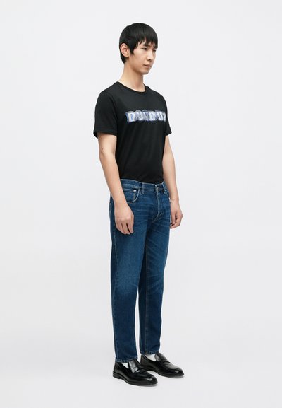 T-shirt noir avec le graphisme "DON'D UP", jean en denim bleu et mocassins noirs. La tenue présente une silhouette simple et ajustée.
