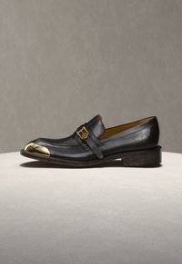 Μαύρο δερμάτινο μοκασίνι με χρυσή μεταλλική επένδυση στη μύτη και διακοσμητικό χρυσό αγκράφα στον ιμάντα, τοποθετημένο σε ουδέτερο γκρι φόντο.