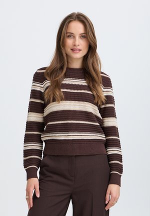 Femme aux longs cheveux bruns portant un pull en tricot rayé marron et beige et un pantalon marron foncé, debout devant un fond uni.