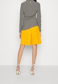 Černobílé kostkované tričko s obtaženým pasem, doplněné o jasně žluté plisované šortky. Modelka má na sobě bílé sandály na tenkých podpatcích.