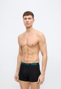 Ευθυτενής νεαρός άνδρας που στέκεται, φορά μαύρα μποξεράκια Puma με μπλε λογότυπο στη ζώνη, ουδέτερο φόντο, τα χέρια χαλαρά στο πλάι.