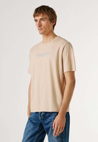 Joven con cabello corto castaño claro, vistiendo una camiseta beige de Pepe Jeans London y jeans azules, posando contra un fondo claro y liso.