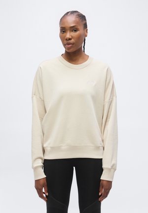 Femme aux cheveux tressés portant un sweat-shirt beige ample et des leggings noirs, debout devant un fond blanc uni.