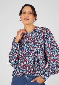 Blusa de manga larga en azul marino con un estampado floral en rojo, rosa y verde. Presenta pliegues y cierre de botones en la parte delantera. Textura de tejido suave.