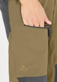 Les pantalons de plein air pour hommes présentent un mélange de tissu vert olive et gris foncé, avec une poche zippée et un logo texturé sur la cuisse.
