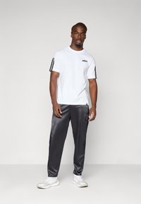 Witte katoenen t-shirt met zwart logo en drie zwarte strepen op de mouwen, gecombineerd met grijze satijnen broek en witte sport schoenen.