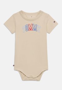 Μπεζ κορμάκι με κοντά μανίκια φτιαγμένο από βαμβάκι, με τη λέξη «TOMMY» σε κόκκινα και μπλε γράμματα. Κουμπώματα πάνας στο κάτω μέρος για εύκολο ντύσιμο.