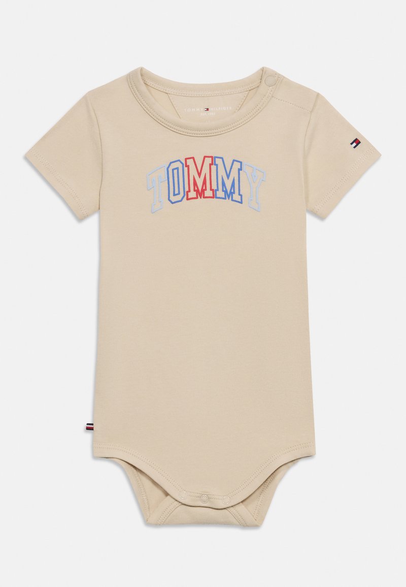 Μπεζ κορμάκι με κοντά μανίκια φτιαγμένο από βαμβάκι, με τη λέξη «TOMMY» σε κόκκινα και μπλε γράμματα. Κουμπώματα πάνας στο κάτω μέρος για εύκολο ντύσιμο.