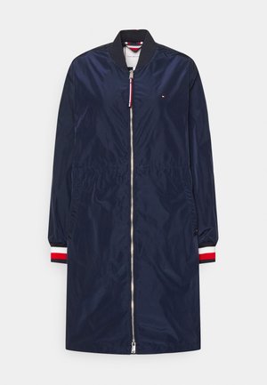 Donkerblauw lange bomberjas met een ritssluiting aan de voorkant, een geribbelde kraag en manchetten met rode, witte en zwarte strepen, en zijzakken.