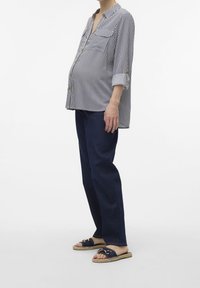 Gravid person i en stribet skjorte med knapper og mørke jeans, stående iført åbne slip-on sandaler mod en ensfarvet hvid baggrund.