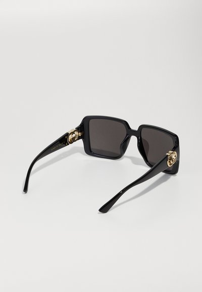 Zwarte vierkante zonnebril met donkere glazen en gouden decoratieve accenten op de pootjes, getoond vanuit een schuine achteraanzicht.