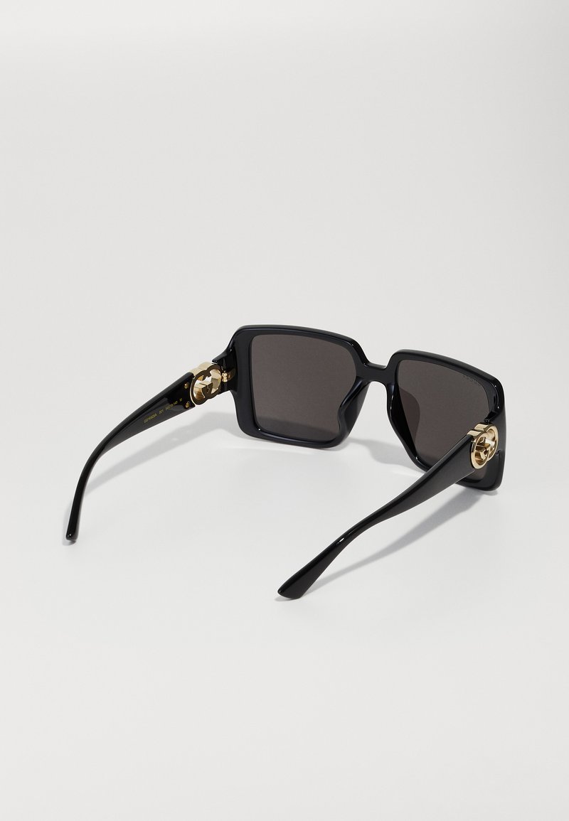 Schwarze quadratische Sonnenbrille mit dunklen Gläsern und goldenen dekorativen Akzenten an den Bügeln, aus einem schrägen hinteren Blickwinkel gezeigt.