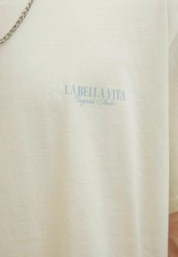 Bežinis medvilnės marškinėliai su užrašu "LA BELLA VITA Originals Studio" šviesiai mėlyna spalva. Apvali iškirptė, trumpomis rankovėmis ir minkšta tekstūra.