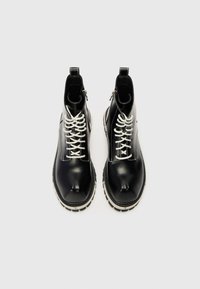 Melnas potītes ar spīdīgu materiālu, ar baltām šņorēm, izteiktu norādītu purngalu un biezu baltu zolīti ar rakstu.