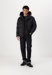 Zwarte pufferjas met een capuchon, voorzien van een gestikt patroon en een knoopsluiting aan de voorkant. Gemaakt over een donker shirt en gecombineerd met donkere denim.