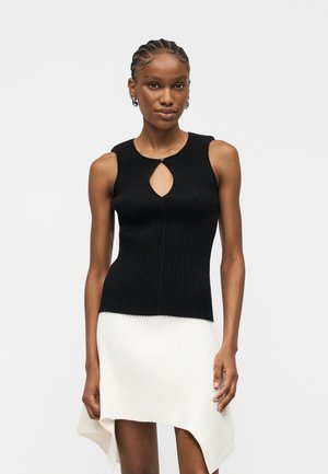 Kobieta z zaplecionymi włosami, ubrana w czarny, bezrękawnik z prążkowanego materiału z ozdobnym wycięciem oraz białą, asymetryczną, prążkowaną spódnicę.