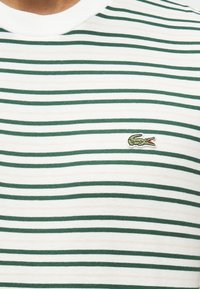 Λευκό και μπεζ πουκάμισο με λεπτές πράσινες οριζόντιες ρίγες και μικρό πράσινο λογότυπο κροκόδειλου κ εντημένο στο στήθος.