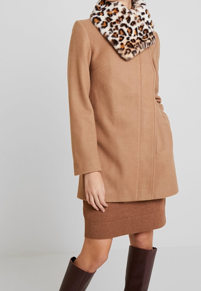 Femme portant un manteau camel avec un col en fausse fourrure à motif léopard, une jupe marron et des bottes hautes marron foncé.