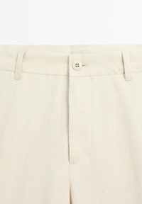 Pantalon beige avec devant plat, fermeture à bouton et passants pour ceinture. Confectionné dans un tissu texturé, avec une poche unique sur le côté gauche.