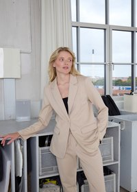 Blonde woman wearing beige suit stands with one hand on a gray shelf in a bright room with large windows and industrial decor.