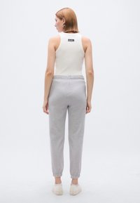 Balts tanktops ar birku aizmugurē, kopā ar gaisīgi pelēkām sporta biksēm ar potīšu manžetēm. Modelis valkā bēšas kurpes.