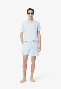 Lichtblauwe poloshirt en bijpassende zwembroek met elastische tailleband. Lacoste-logo op beide items. Model draagt een zonnebril, blootvoets.