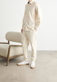 Camicia beige a maniche lunghe con bottoni, pantaloni beige coordinati e sneakers bianche. Una sedia rivestita di un colore chiaro è nelle vicinanze.