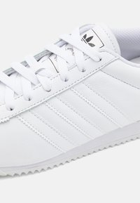 Bílá kožená teniska s třemi texturovanými proužky, kulatou špičkou, plochými bílými tkaničkami a vroubkovanou gumovou podrážkou pro lepší trakci.