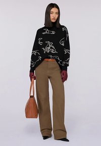 Černý svetr s bílým potiskem psa, béžové kalhoty s vysokým pasem, vínové rukavice a hnědá kabelka. Modelka předvádí moderní, ležérní outfit.