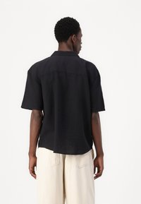 Camisa negra de manga corta, confeccionada en lino, con un corte relajado, que presenta un cuello con puntas y un dobladillo ligeramente curvado.