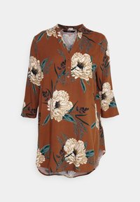 Brązowa tunika w kwiaty z dużymi kremowymi kwiatami, zielonymi liśćmi, rękawami 3/4, zaokrąglonym dołem i guzikiem przy dekolcie w kształcie litery V.