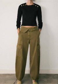 Pull court noir côtelé avec des boutons aux épaules, associé à un pantalon cargo ample vert olive avec poches latérales et poignets élastiques.