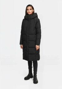 Abrigo acolchado negro con un cuello alto, bolsillos con cremallera y un diseño hasta las rodillas, que presenta un patrón acolchado y una capucha desmontable.