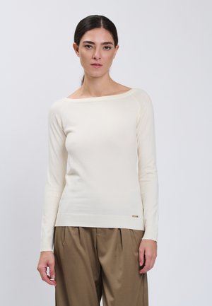 Kvinde med mørkt hår i lav hestehale bærer en cremefarvet langærmet sweater og løse brune bukser mod en lysegrå baggrund.