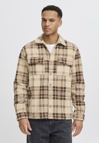 Beige and brown plaid flannel shirt with a spread collar, front button closure, and two chest pockets. Soft texture and relaxed fit.