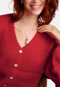 Roter, flauschiger Cardigan mit tiefem V-Ausschnitt und weißen Knöpfen. Eine silberne Halskette mit einem bunten Anhänger und einer Perle hängt am Ausschnitt.