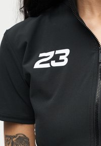 Μαύρο πουκάμισο με φερμουάρ και λευκό "23" στο αριστερό στήθος και ορατό τατουάζ στο χέρι κάτω από το κοντό μανίκι.