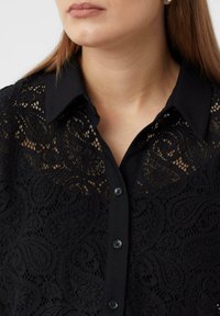 Black blouse featuring a lace design with intricate patterns. Collared with button closure, displaying a smooth texture and sheer accents.