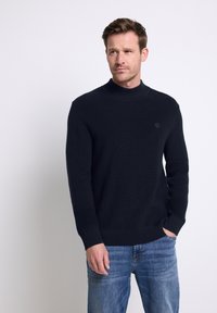 Pull en maille bleu marine foncé avec un col montant, texture côtelée et un logo subtil. Porté avec un jean en denim bleu clair.