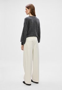 Γκρι πλεκτό πουλόβερ με μακριά μανίκια, συνδυασμένο με φαρδιά λευκά παντελόνια και μαύρα παπούτσια. Το μοντέλο απεικονίζεται από πίσω, σε μινιμαλιστικό φόντο.